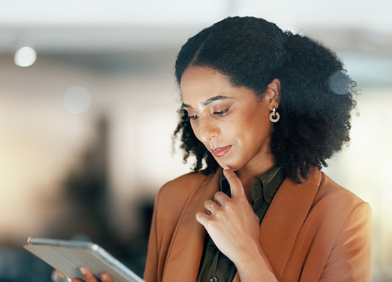 une femme professionnelle avec des cheveux bouclés, portant un blazer beige et des boucles d'oreilles dorées. Elle regarde attentivement une tablette, le fond flou avec des lumières bokeh crée une ambiance chaleureuse et moderne.
